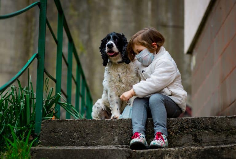 A littler girl wearing a mask sitting on the steps outside their house with her dog during the 2019 Coronavirus pandemic
